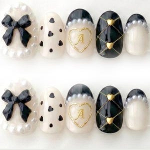 May include: A set of press-on nails with various designs. One nail features a black bow, others have black hearts, a gold heart with the letter "A", and a black grid pattern with gold hearts. The nails are primarily white, black, and gold.