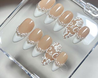 May include: A set of ten almond-shaped press-on nails featuring a nude base and white tips. Most nails are accented with a row of small white pearls along the smile line. Two nails feature white embossed floral patterns and the text C&C.