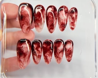 May include: A set of ten almond-shaped press-on nails featuring a glossy finish with a marbled pattern in deep burgundy, dark red, and white swirls. The nails are arranged in two rows inside a clear, rectangular plastic storage container.