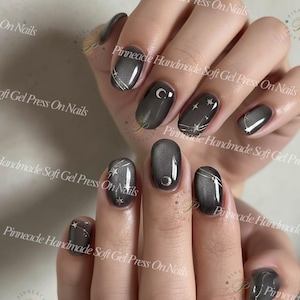 May include: Dark grey soft gel press-on nails with silver celestial designs. Some nails feature a crescent moon or stars and lines. The text "Pinneade Handmade Soft Gel Press On Nails" is visible.