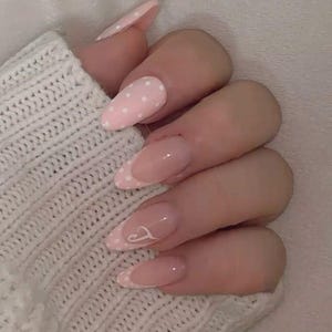 Puede incluir: Primer plano de una mano con uñas largas en forma de almendra. Algunas uñas están pintadas de rosa claro con lunares blancos, mientras que otras tienen un diseño de manicura francesa con un pequeño corazón blanco. Las uñas están bien cuidadas y brillantes.