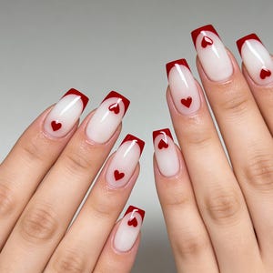Può includere: Unghie lunghe e squadrate con base bianca e punta francese rossa vibrante. Ogni unghia presenta un piccolo design a cuore rosso, creando un look romantico ed elegante. Le unghie sono ben curate e mostrano una finitura pulita e lucida.