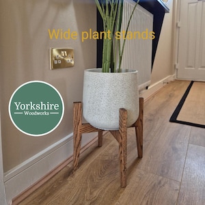 May include: A speckled grey plant pot with a tall green plant, on a wooden plant stand. The stand has a mid-century modern design with four angled legs. The text "Wide plant stands" is visible.