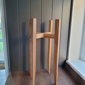 May include: A wooden plant stand with a simple, rectangular design. The stand is made of light brown wood and has four legs.