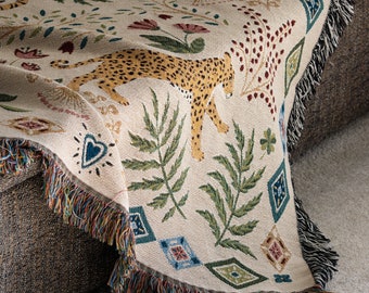 Peut inclure: Plaid beige avec un motif répété de léopard, de fleurs et de feuillage. La couverture a un bord à franges multicolores. Le motif comprend un léopard avec des taches jaunes, des feuilles vertes et des fleurs rouges et roses.