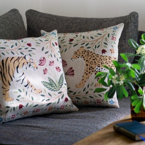 May include: Two decorative cushions with animal and floral designs. One cushion features a tiger, the other a leopard, both surrounded by colourful flowers and foliage. The cushions are on a grey sofa.