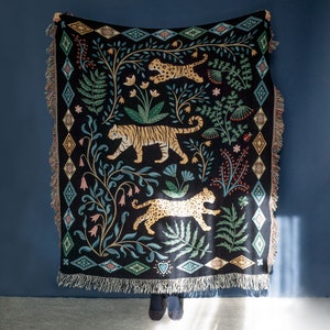 May include: Black woven throw blanket with a jungle-themed design. The blanket features tigers and leopards, along with floral and geometric patterns in shades of green, blue, and tan. The edges have a fringed trim.