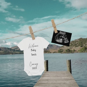 May include: A white onesie with the text "Welcome Baby Smith Coming 2024" hanging on a clothesline with a black and white ultrasound image hanging next to it. The onesie and ultrasound image are hanging over a body of water with a wooden dock in the foreground.