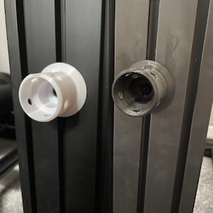 以下が含まれることがあります： 中央に円形の開口部のある、白と黒の2つの3Dプリントされたプラスチック部品。部品は黒い表面に取り付けられています。