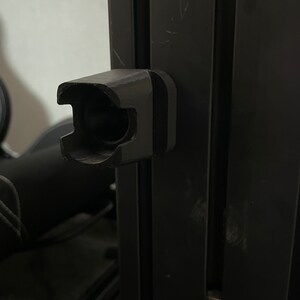 Op de afbeelding: Een zwarte kunststof beugel met een rond gat in het midden, bevestigd aan een zwart metalen frame.