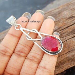Puede incluir: Colgante de plata con una gran piedra preciosa de rubí de talla ovalada. El rubí es de color rojo intenso y está engastado en un marco de plata. El colgante tiene un diseño geométrico abierto y el texto "SILVERJEWELS92".