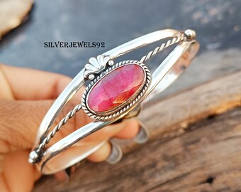 Puede incluir: Un brazalete de plata con una piedra preciosa roja engastada en un bisel ovalado de plata. El brazalete tiene un diseño de alambre trenzado y un acento floral decorativo.