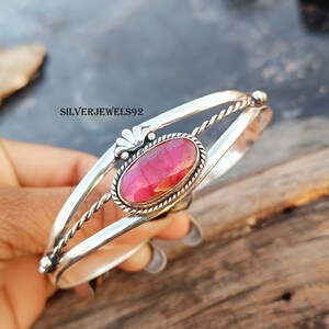 May include: A silver bangle bracelet featuring a red gemstone set in a silver oval bezel. The bracelet has a twisted wire design and a decorative floral accent.