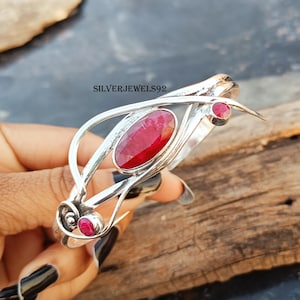 May include: A silver bangle bracelet with a delicate, intricate design featuring three red ruby gemstones. The bracelet is open and adjustable.