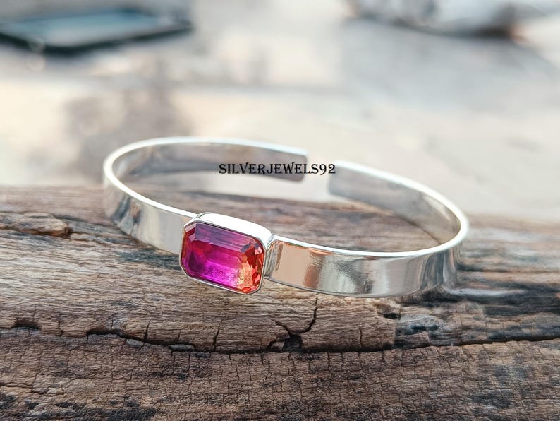 May include: A silver bangle bracelet with a pink and orange rectangular gemstone. The bracelet is on a wooden surface. SILVERJEWELS92 is printed on the bracelet.