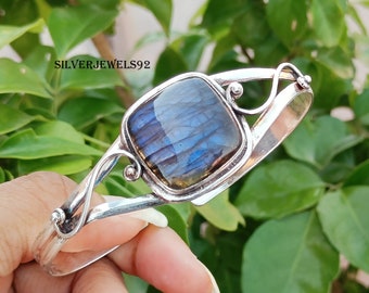 Puede incluir: Una pulsera de plata con una piedra de labradorita cuadrada e iridiscente en el centro. La piedra muestra tonos azules y morados. La pulsera tiene un diseño calado y el texto "SILVERJEWELS92".
