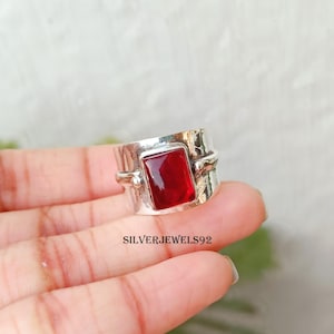 May include: A wide silver band ring featuring a rectangular, deep red gemstone set in the center. The silver metal has a textured, hammered finish with small spherical accents on either side of the stone. The text SILVERJEWELS92 is visible.
