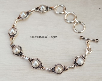 May include: A silver bracelet with a toggle clasp features a chain with seven white pearl accents. Each pearl is set in a silver bezel with a decorative border.