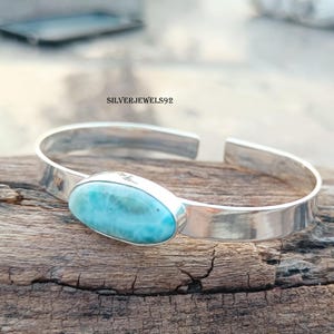 May include: A silver bangle bracelet featuring a light blue oval-shaped gemstone. The gemstone has a distinctive iridescent quality.