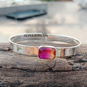 May include: A silver bangle bracelet with a pink and orange gemstone set in the center. The bracelet is on a wooden surface.
