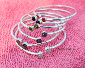 May include: A set of six silver bangle bracelets with different colored gemstones. Each bracelet has a single gemstone set in a silver bezel. The gemstones are red, black, green, purple, brown, and white.