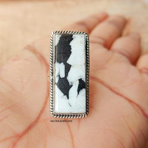 May include: A rectangular ring with a white and black stone set in a silver-colored metal. The stone has a unique pattern of black and white, and the ring has a twisted rope-like border. The text "SILVERJEWELS92" is visible.