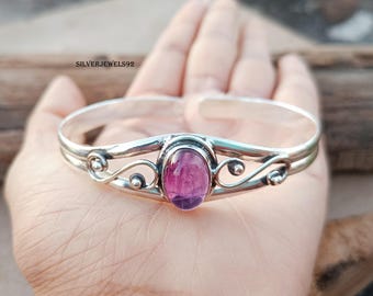 May include: A silver-toned cuff bracelet with an oval, magenta-colored gemstone at the center. The bracelet features decorative swirls and a polished finish. The text "SILVERJEWELS92" is visible.