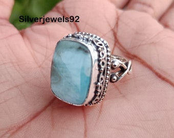 Könnte beinhalten: Ein silberner Ring mit einem hellblauen Edelstein, der in einer quadratischen Fassung sitzt. Der Ring hat ein dekoratives Band mit einem floralen Design.