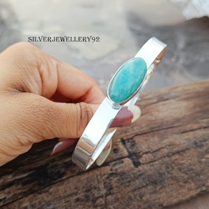 May include: A silver bangle bracelet with a light green oval gemstone. The bracelet is open and has a smooth, polished finish.