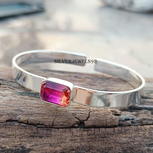 May include: A silver bangle bracelet with a pink and orange rectangular gemstone. The bracelet is on a wooden surface. SILVERJEWELS92 is printed on the bracelet.