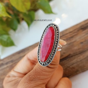 May include: A silver ring with a large oval-shaped red gemstone set in a decorative silver bezel. The ring is on a finger.