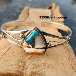 May include: A silver bangle bracelet with a triangular turquoise and white stone set in the center. The bracelet has a delicate, twisted design.