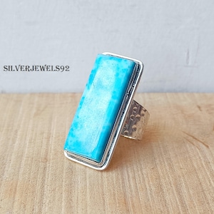 May include: A silver ring with a rectangular turquoise gemstone. The light blue gemstone is set in a silver frame. The band has a hammered texture. The text "SILVERJEWELS92" is visible. The ring is a statement piece.