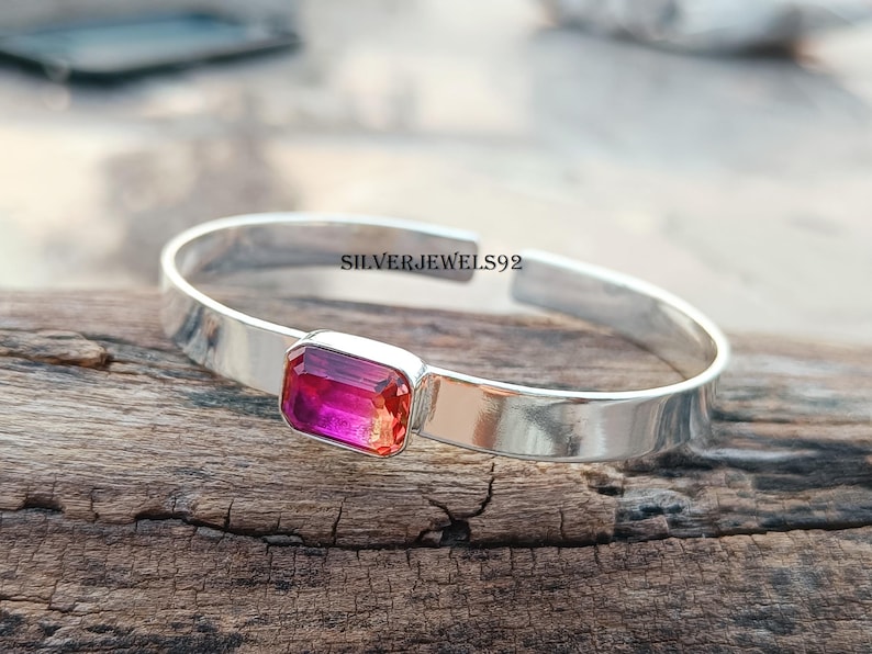 May include: A silver bangle bracelet with a pink and orange rectangular gemstone. The bracelet is on a wooden surface. SILVERJEWELS92 is printed on the bracelet.