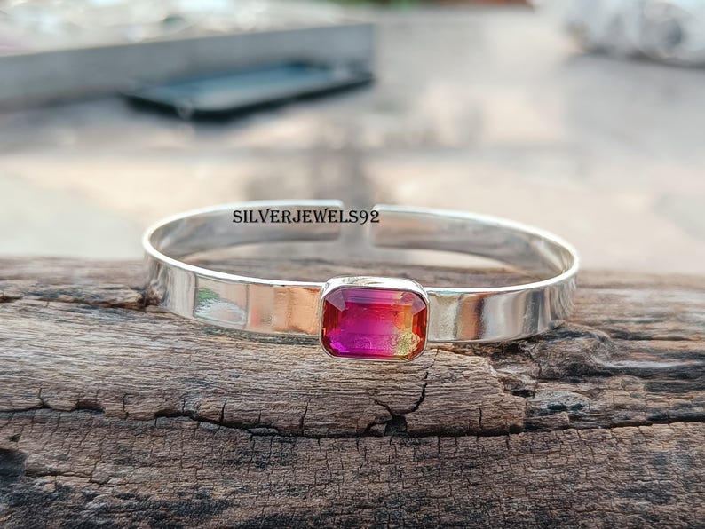 May include: A silver bangle bracelet with a pink and orange gemstone set in the center. The bracelet is on a wooden surface.