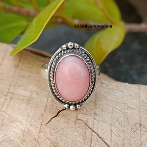 May include: A silver ring with an oval, light pink gemstone. The gemstone is set in a detailed, ornate silver frame with a twisted rope design around the edge. The ring is on a wooden surface.