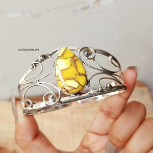 May include: A silver cuff bracelet with an openwork design and a large, teardrop-shaped yellow stone with brown veining. The bracelet is held up by a hand.