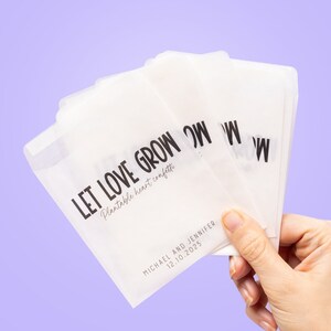 May include: Several translucent white packets, each printed with the words "LET LOVE GROW" in black, and "Plantable heart confetti". The packets also display the names "MICHAEL AND JENNIFER" and the date "12.10.2025". The background is a pale purple.