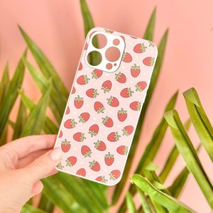 Puede incluir: Funda de teléfono con un patrón repetido de fresas rojas sobre un fondo rosa claro. Las fresas tienen tallos verdes y están dispersas por toda la funda. La funda tiene un borde blanco y recortes para la cámara y los botones del teléfono.