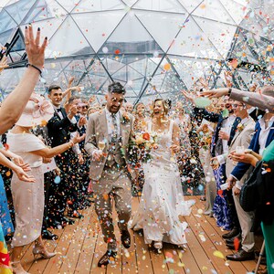 May include: A bride and groom walk through a confetti shower as guests cheer and throw confetti. The couple is walking through a glass structure with a wooden floor.