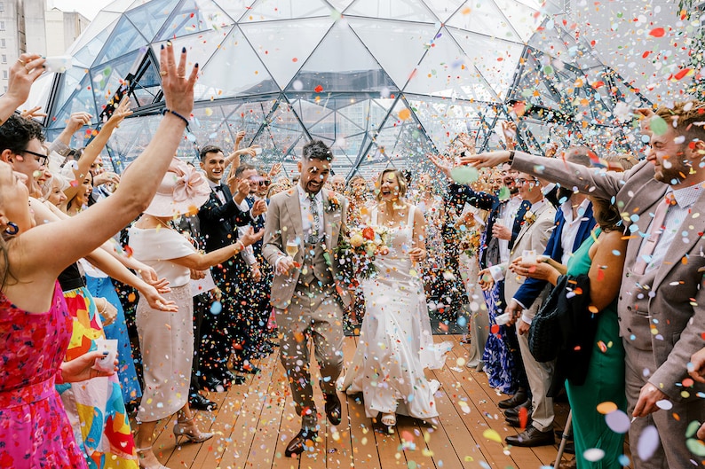 May include: A bride and groom walk through a confetti shower as guests cheer and throw confetti. The couple is walking through a glass structure with a wooden floor.