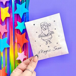 以下が含まれることがあります： サンタクロースと星の黒い線画が描かれた、小さなオフホワイトの紙袋。「Magic Snow」の文字が筆記体で書かれています。背景にはカラフルな星の飾りとストリーマーがあります。