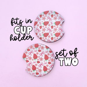 May include: Set of two white ceramic cup coasters with a pink and red strawberry and bow pattern. The text "fits in cup holder" and "set of two" is printed in black.