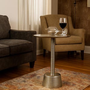 May include: A small, round, metallic side table with a ribbed base and stem. A wine glass with red wine and an empty wine glass rest on the table's surface. The table sits on a patterned rug in a living room setting.