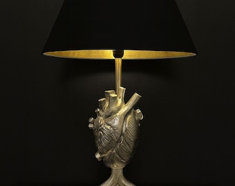 Puede incluir: Lámpara de mesa única con un diseño detallado de corazón anatómico en acabado metálico. La lámpara tiene una pantalla negra con interior dorado y una base cuadrada. La lámpara está sobre una superficie oscura.