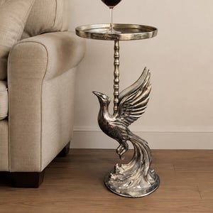 May include: A silver-toned metal side table featuring a circular top supported by a central column and a base sculpted in the shape of a bird with outstretched wings. A clear glass stemware filled with red liquid sits on the table surface.
