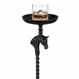 May include: A matte black pedestal side table featuring a circular top, a central support column with a twisted rope design, and a sculpted horse head detail. A clear glass tumbler containing an amber liquid and ice cubes sits on the table top.