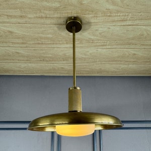 Może przedstawiać: Wisząca lampa z mosiądzu z okrągłym, płaskim kloszem i białym, matowym dyfuzorem szklanym. Lampa jest zawieszona na suficie z drewnianymi panelami.