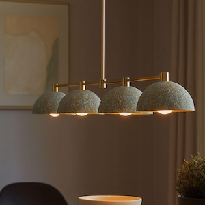 May include: A modern, four-light pendant lamp with a brass finish. The lampshades are a speckled, dome-shaped design in shades of blue and gold. The lamp hangs above a wooden table, with a bowl and plant visible.