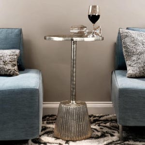May include: A silver side table with a round top and a fluted base. Two wine glasses, one filled with red wine, rest on the table. The table is positioned between two blue armchairs with patterned pillows. The rug has a black and white pattern.